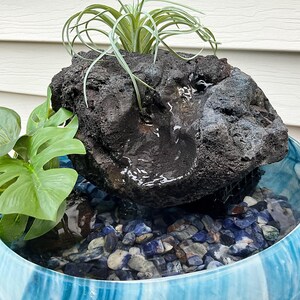 May include: A decorative water fountain featuring a dark, textured rock formation with a small waterfall. The fountain sits in a blue and white glass bowl filled with blue and gray stones. Green plants add a natural touch.