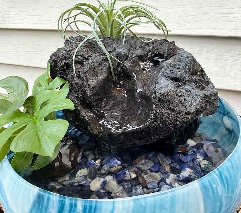 May include: A decorative water fountain featuring a dark, textured rock formation with a small waterfall. The fountain sits in a blue and white glass bowl filled with blue and gray stones. Green plants add a natural touch.