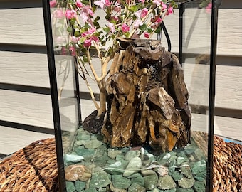 May include: A glass terrarium with a black metal frame, featuring a miniature landscape. Inside, there's a rock formation, green pebbles, and a small tree with pink flowers. The terrarium sits on a woven surface.
