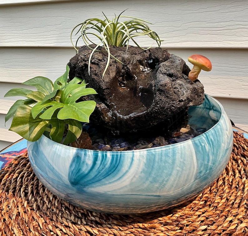 May include: A decorative indoor water fountain featuring a dark, textured rock formation with a small mushroom accent. The rock sits in a blue and white swirled glass bowl, with green plants and a small air plant. The bowl rests on a woven mat.