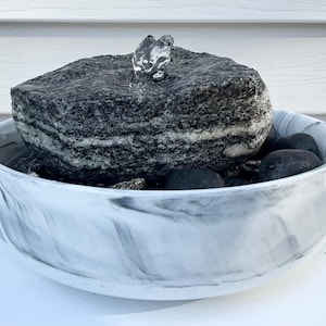 Op de afbeelding: Een kleine, decoratieve waterfontein. Een donkergrijze, gelaagde rots zit in een wit en grijs gemarmerde kom. Water stroomt uit de bovenkant van de rots. Zwarte stenen bevinden zich aan de basis van de rots.
