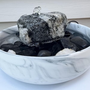 Op de afbeelding: Een kleine fontein voor binnenshuis met een zwart-witte granieten rots. De rots staat in een witte kom met marmerpatroon, gevuld met water en zwarte kiezels. Een kleine waterstraal stroomt uit de bovenkant van de rots.