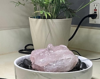 May include: A decorative water fountain featuring a large, light pink rose quartz stone. The stone sits atop black river rocks in a white marble-patterned bowl. A potted plant is in the background.