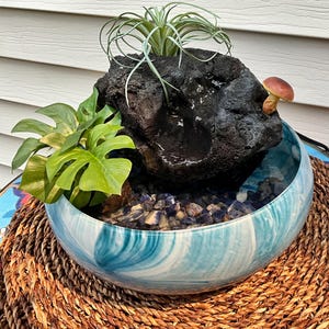 May include: A decorative indoor water fountain. A dark, porous rock formation sits atop a blue and white ceramic bowl. The bowl contains water and small blue and gray stones. Green plants and a small mushroom add detail.