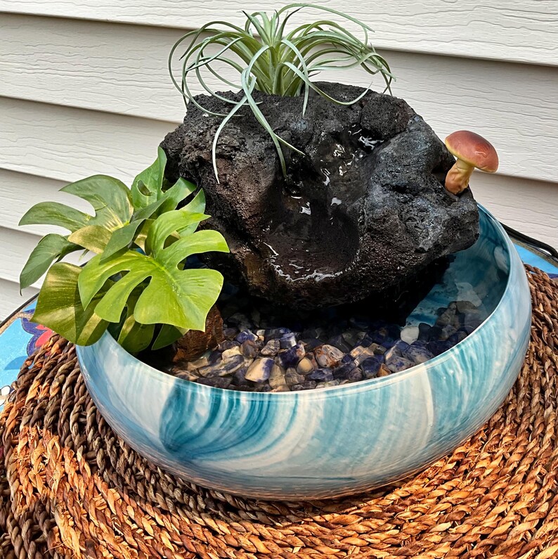 May include: A decorative indoor water fountain. A dark, porous rock formation sits atop a blue and white ceramic bowl. The bowl contains water and small blue and gray stones. Green plants and a small mushroom add detail.