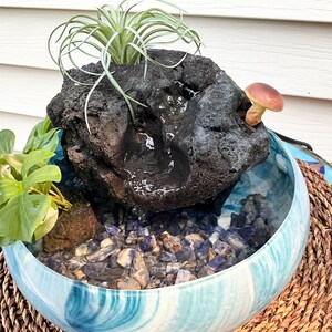 May include: A decorative indoor water fountain featuring a dark, textured rock formation with a small waterfall. The fountain sits in a blue and white swirled ceramic bowl filled with blue and gray stones. A small mushroom and plants are included.