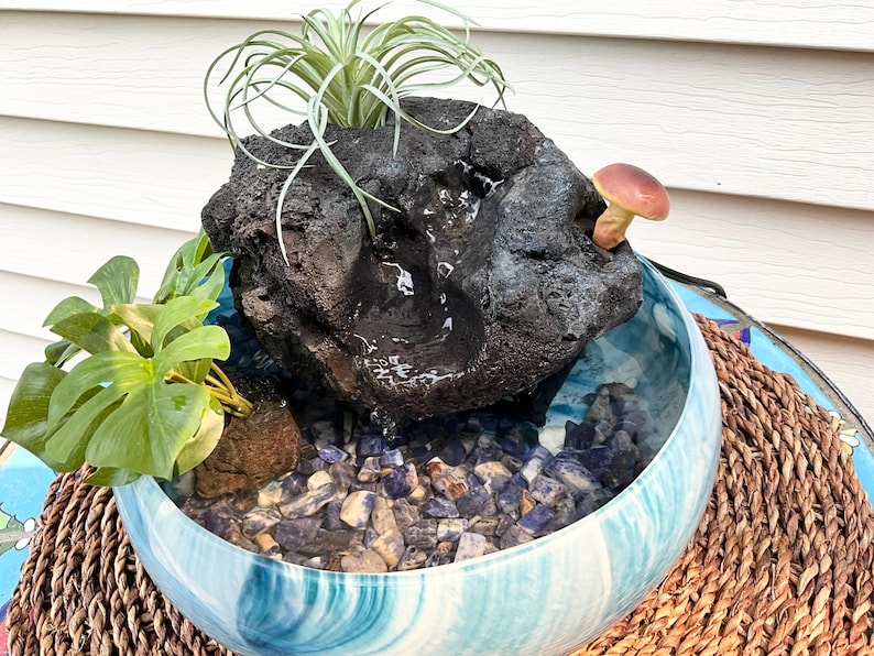 May include: A decorative indoor water fountain featuring a dark, textured rock formation with a small waterfall. The fountain sits in a blue and white swirled ceramic bowl filled with blue and gray stones. A small mushroom and plants are included.
