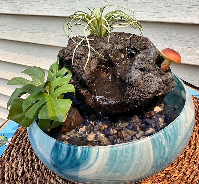 May include: A decorative indoor water fountain with a natural rock feature, a small mushroom, and lush green plants. The fountain sits in a blue and white swirled ceramic bowl filled with small stones.
