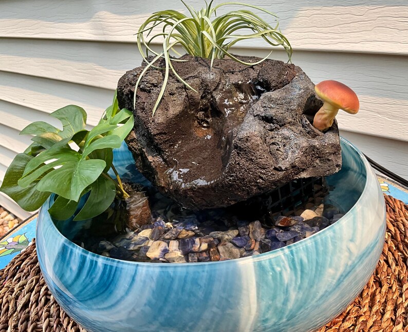 May include: A decorative water fountain with a blue and white marbled bowl. A dark, textured rock sits in the center, with a small mushroom and greenery. The bowl is filled with water and small stones.