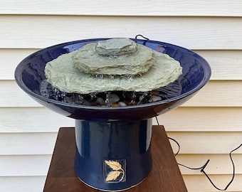 May include: A blue ceramic tabletop fountain with a three-tiered rock waterfall. The fountain has a leaf design on the base and sits on a wooden pedestal.
