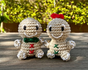 May include: Two crocheted gingerbread people, one with a red bow and the other with a green bow, are standing side by side on a wooden surface.