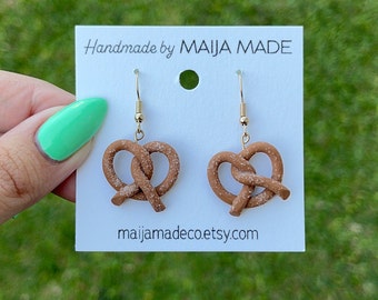 Pode incluir: Brincos de pretzel feitos à mão com ganchos dourados. Os brincos castanhos têm uma leve camada de pó branco, que simula o sal. São exibidos em um cartão branco com o texto "Handmade by MAIJA MADE" e o endereço do site.