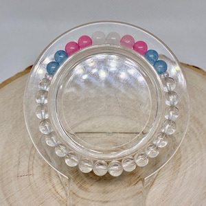 Peut inclure: Un bracelet composé de perles sphériques transparentes, bleu clair, roses et blanches disposées en cercle. Le bracelet est présenté sur un support circulaire transparent, mettant en valeur l'agencement des perles translucides et opaques.