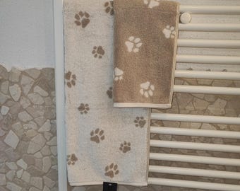 May include: Two towels hanging on a white towel rack. The top towel is brown with white paw prints. The bottom towel is white with brown paw prints.