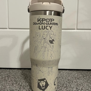 Può includere: Un bicchiere termico beige con coperchio e manico color crema. Il bicchiere presenta un'illustrazione in bianco e nero di tre donne e altre grafiche, con il testo "KPOP DEMON HUNTERS LUCY".