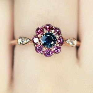 Könnte beinhalten: Ein blütenförmiger Ring mit einem dunkelblauen zentralen Edelstein, umgeben von kleineren rosa und lila Steinen. Der Ring hat ein roségoldenes Band und birnenförmige Akzente auf beiden Seiten des Blumendesigns.