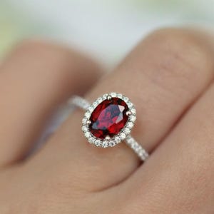 Könnte beinhalten: Ein silberner Ring mit einem großen, oval geschliffenen, tiefroten Edelstein, umgeben von einem Kranz aus kleineren, klaren Steinen. Das Ringband ist ebenfalls mit kleinen, funkelnden Steinen besetzt.