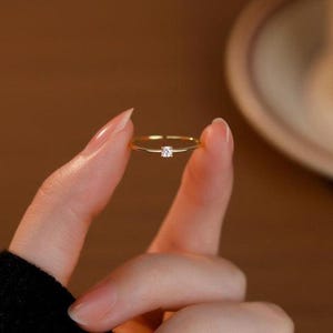 May include: A slender gold ring featuring a small, square-cut diamond. The ring is held between fingers, highlighting its minimalist design and elegant simplicity. The band is thin and polished, reflecting light. A classic piece of jewellery.