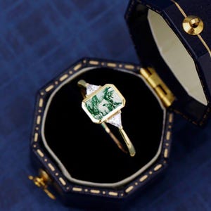 May include: A gold ring with a rectangular green gemstone, flanked by two triangular white gemstones. The ring is presented in a dark blue jewelry box with a gold clasp.