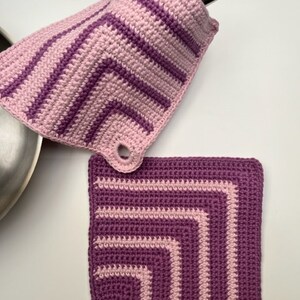 Puede incluir: Dos agarraderas de olla de ganchillo en rosa y morado con un patrón a rayas. Las agarraderas tienen un lazo para colgar.