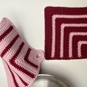 Puede incluir: Dos agarraderas de olla de ganchillo en rosa y burdeos. Una es una agarradera de olla cuadrada y la otra es una agarradera de olla colgante.