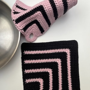 Puede incluir: Dos agarraderas de olla de crochet en negro y rosa con un patrón de chevron. Una agarradera de olla es de forma cuadrada y la otra es de forma larga y estrecha.