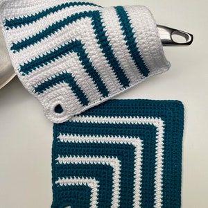 Puede incluir: Dos agarraderas de olla de ganchillo con un patrón de rayas blancas y azul verdoso. Las agarraderas tienen un agujero en la esquina para colgarlas.