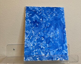 May include: A blue abstract watercolor painting on canvas with a white background. The painting is displayed on a clear acrylic shelf.