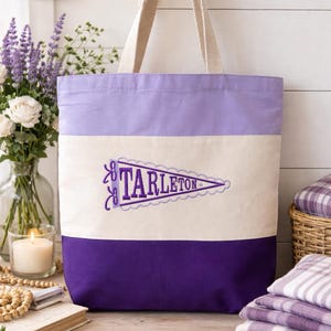 May include: A canvas tote bag with three horizontal stripes in purple, cream, and lavender. The cream stripe features an embroidered pennant with the word "TARLETON" in purple. The bag has beige handles.