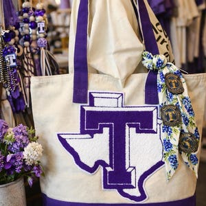 May include: Cream-colored tote bag with purple accents. Features a large purple and white Texas-shaped logo with a letter "T". The bag has purple handles and a purple bottom. A floral scarf with a matching logo is tied to the handle. Other purple accessories are visible.
