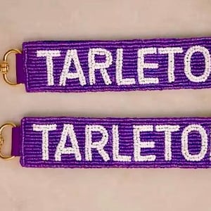 May include: Two purple keychains with the word "TARLETON" in white beads. Each keychain has a gold-colored clasp. The keychains are rectangular and appear to be made of woven material.