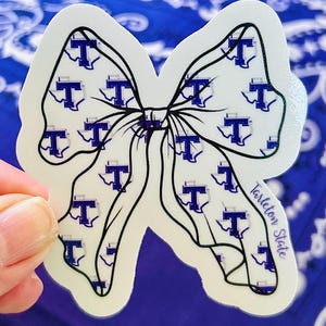 May include: A white sticker in the shape of a bow with a black outline. The bow is patterned with blue "T" logos inside of the state of Texas. The text "Tarleton State" is printed on the right side of the bow.