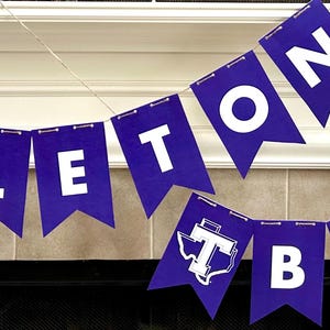 May include: A purple banner with white lettering that reads "TARLETON BOUND" is strung across a fireplace. The banner features pennant-shaped flags with the university's logo. The banner is a festive decoration.