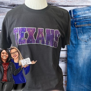 May include: A dark gray t-shirt with the word "TEXANS" in sparkling purple and silver rhinestones. The shirt is displayed with a pair of blue jeans. Two cartoon women are holding accessories.