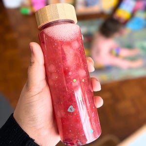 May include: A clear sensory bottle filled with red liquid, glitter, and small shapes. The bottle has a bamboo lid and is held in a hand. The liquid has bubbles at the top. The bottle is a children's toy.