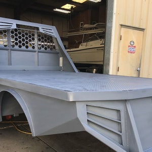 Puede incluir: Una plataforma de camión plana de metal gris con una jaula de metal con patrón de panal detrás de la cabina. La plataforma tiene una superficie de placa de diamante y un parachoques trasero con un enganche.