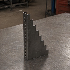 Può includere: Uno strumento a gradino in metallo grigio scuro con una serie di fori praticati lungo un lato. Lo strumento ha un design a gradini su un bordo e una piegatura ad angolo retto nella parte superiore. Strumento per la lavorazione dei metalli.