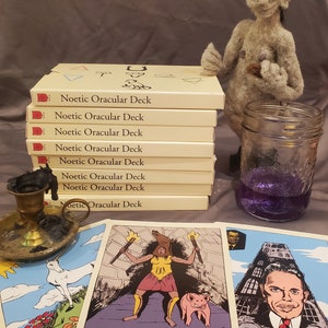 May include: A stack of eight cream-colored boxes with the text "Noetic Oracular Deck" printed on each box. The boxes are stacked on a gray surface. There are three cards from the deck on the surface, one with the text "FURY" and another with the text "Iron Pris".
