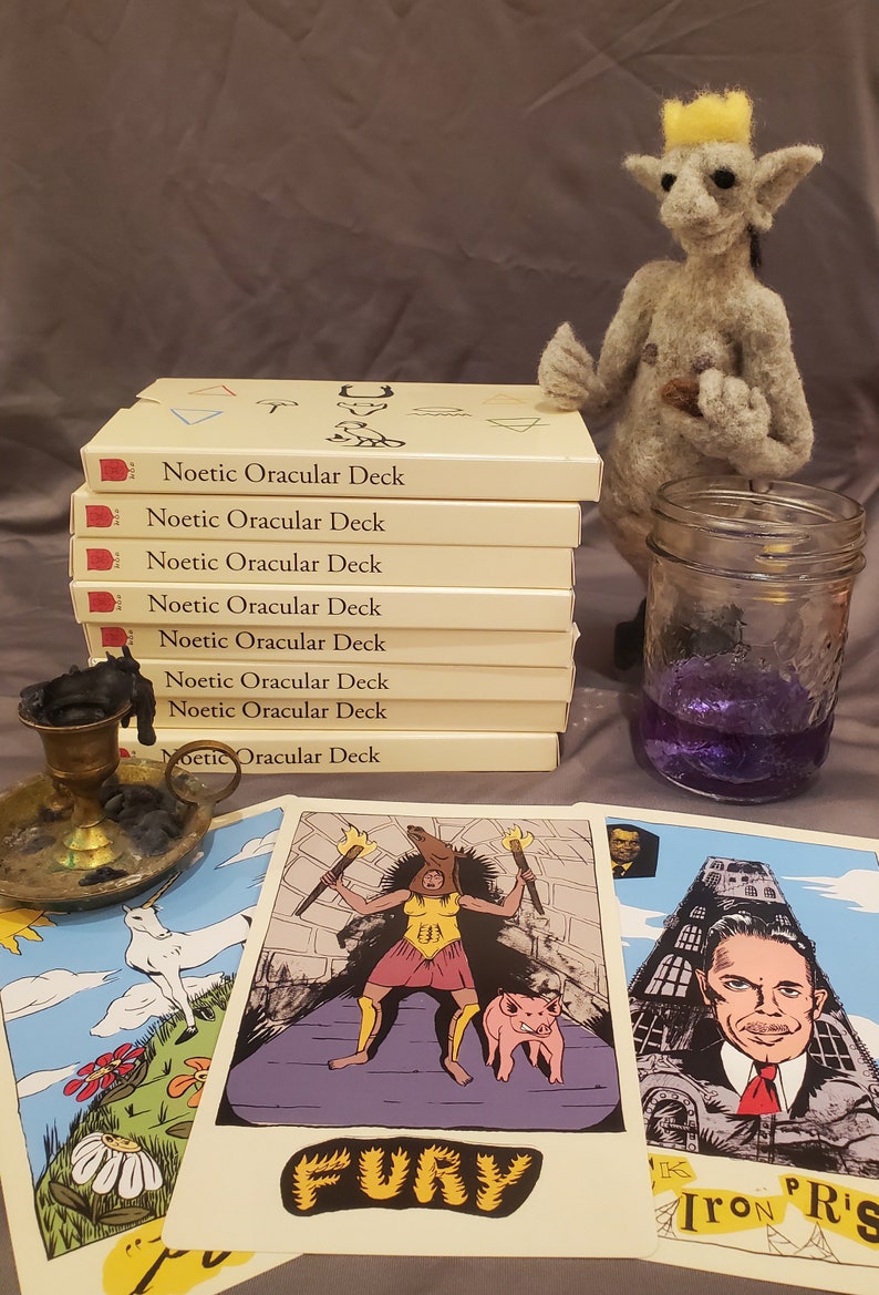 May include: A stack of eight cream-colored boxes with the text "Noetic Oracular Deck" printed on each box. The boxes are stacked on a gray surface. There are three cards from the deck on the surface, one with the text "FURY" and another with the text "Iron Pris".