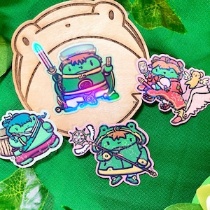 May include: A collection of colorful stickers featuring frog characters in various poses. One frog holds a sword, another a staff, and others are depicted with accessories. The stickers are on a green background with wooden frog-shaped base.