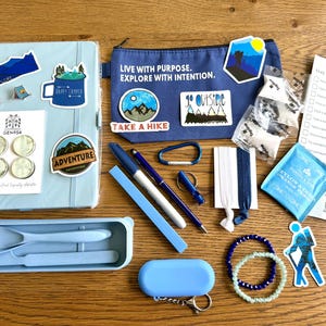 May include: A light blue notebook with adventure stickers, a blue pencil case with the text LIVE WITH PURPOSE, EXPLORE WITH INTENTION, a blue cutlery set, two pens, a carabiner, a whistle, hair ties, a tea bag, a checklist, and two beaded bracelets.