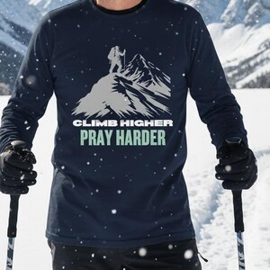 May include: A navy blue long-sleeve shirt with the text "CLIMB HIGHER PRAY HARDER" and a mountain graphic. The shirt is worn by a person in a snowy mountain landscape, wearing a brown beanie and black gloves, holding ski poles.