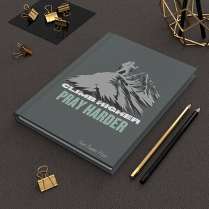 May include: A gray journal with the text "CLIMB HIGHER PRAY HARDER" and a mountain graphic. The journal has a space for a name and is next to gold and black pencils and gold binder clips.