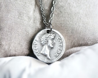 May include: A silver chain necklace with a round silver pendant featuring a profile of a person with the word "CAESAR" inscribed on the pendant.