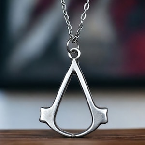 May include: A silver pendant necklace featuring the Assassin's Creed symbol. The pendant is a stylized teardrop shape with a pointed bottom and two small wings on either side.