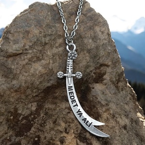 May include: A silver necklace with a sword pendant. The sword is curved and has the words "MEDET YA ALI" inscribed on it.