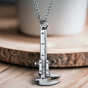 Pode incluir: Um colar de prata com um pingente em forma de calibre de medição. O pingente tem marcas para 0, 1, 2, 3 e 4.