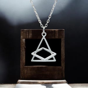 May include: A silver-colored pendant necklace featuring a geometric design. The pendant has a triangular shape with a diamond shape inside. The necklace is displayed on a wooden stand against a dark background.