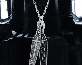 May include: A silver-colored pendant necklace featuring two coffin-shaped charms. One charm has a cross design, and the other depicts a skeleton. The necklace is suspended from a delicate chain, creating a gothic or alternative aesthetic.