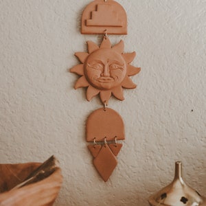 May include: A terracotta sun with a face, geometric shapes, and a hanging loop. The sun is hanging from a string and is attached to a terracotta arch. The arch is attached to a terracotta triangle with a hanging loop. The terracotta pieces are hanging on a white wall.